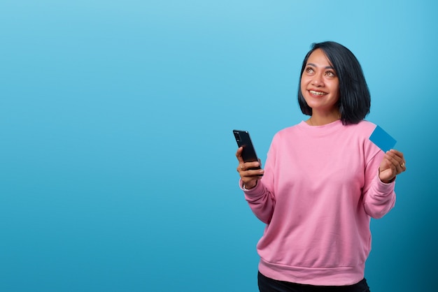 Retrato de hermosa mujer asiática sonriente con teléfono móvil y sosteniendo una tarjeta de crédito y buscando un espacio vacío