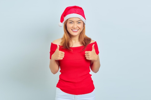 Retrato de hermosa mujer asiática sonriente con vestido de Navidad y mostrando los pulgares hacia arriba o signo de aprobación sobre fondo blanco.