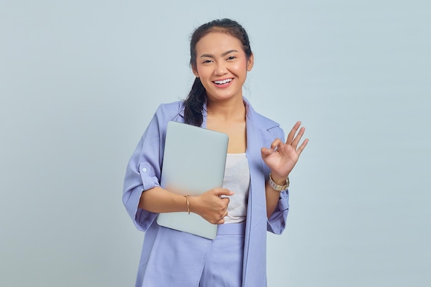 Foto retrato de una hermosa mujer asiática sosteniendo una laptop sonriendo y mostrando un signo de ok sobre fondo blanco