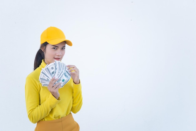 Retrato de una hermosa mujer asiática con telas amarillas en una pared blanca sucia. Las chicas hipster usan un sombrero amarillo y toman una foto. La gente de Tailandia tiene mucho dinero en dólares en la mano.