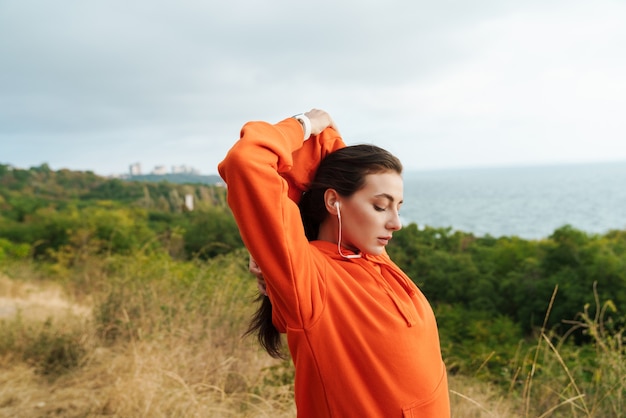 Foto retrato de hermosa mujer atlética en ropa deportiva estirando sus brazos y usando auriculares mientras hace ejercicio en la orilla del mar