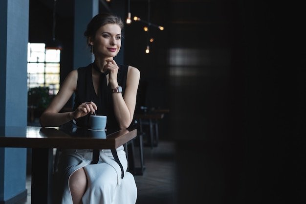 Retrato de hermosa mujer bebiendo té o café y mirando con una sonrisa por la ventana de la cafetería mientras disfruta de su tiempo libre, agradable almuerzo de mujer de negocios en la cafetería moderna durante su descanso laboral.