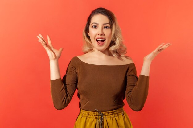 Retrato de hermosa mujer bonita asombrada con cabello rubio en blusa marrón de pie sorprendida con la boca abierta levantó las manos y mirando a la cámara Disparo de estudio interior aislado sobre fondo rojo