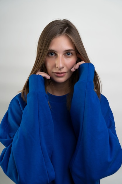 Retrato de una hermosa mujer de cabello castaño con ojos azules vistiendo una sudadera deportiva y posando sobre un fondo blanco.