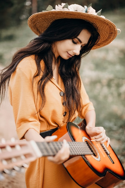 Retrato de una hermosa mujer con cabello largo oscuro cantando en una guitarra afuera en la naturaleza con un sombrero.