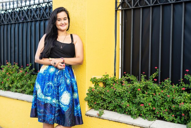 Retrato de una hermosa mujer con cabello negro y vestido negro apoyado contra una pared amarilla