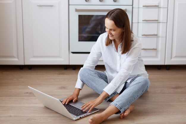 Retrato de hermosa mujer de cabello oscuro sentada en el piso en la cocina, mirando la pantalla del portátil con expresión facial alegre positiva, vestida con camisa blanca y jeans.