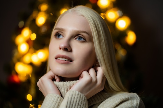 Retrato de una hermosa mujer con cabello rubio cerca de un árbol de Navidad decorado, ella está vestida con un suéter de punto blanco