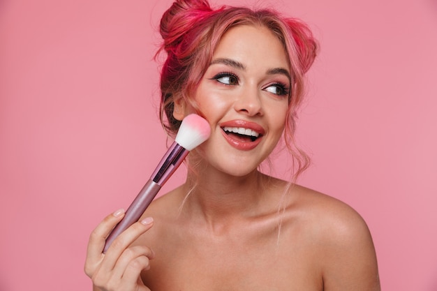 Retrato de hermosa mujer sin camisa con peinado colorido aplicando cosméticos con pincel de maquillaje