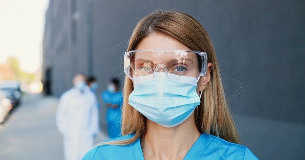 Retrato de hermosa mujer caucásica médico en máscara médica y gafas mirando a la cámara. Cerrar médico femenino en protección respiratoria. Médicos multiétnicos en el fondo. Acercándose. Tiro con plataforma rodante