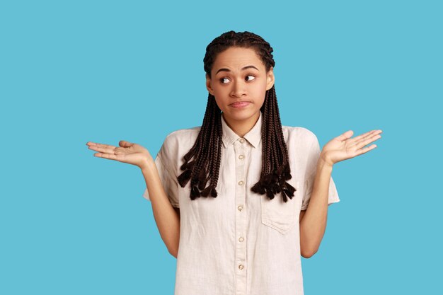 El retrato de una hermosa mujer confundida con rastas negras se siente inseguro porque no puede encontrar el camino correcto, levantando los brazos, usando camisa blanca. Disparo de estudio interior aislado sobre fondo azul.