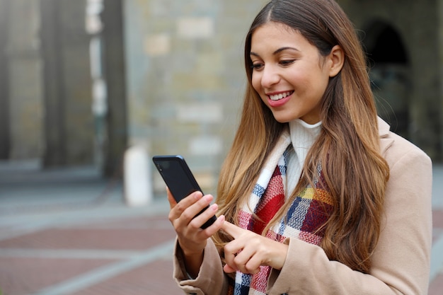 Retrato de hermosa mujer joven con abrigo y bufanda en la ciudad escribiendo en su teléfono