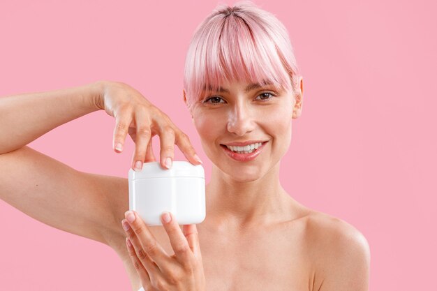 Retrato de hermosa mujer joven con cabello rosado sosteniendo un tarro blanco con loción corporal hidratante después