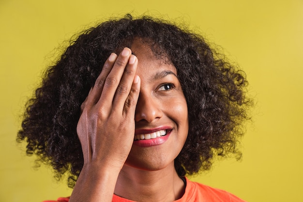 Retrato de hermosa mujer joven con estilo afro de pie sobre fondo amarillo aislado cubriendo un ojo con la mano.