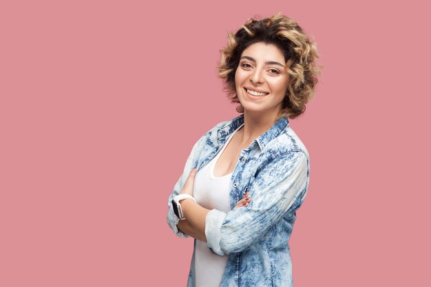 Retrato de hermosa mujer joven feliz exitosa con peinado rizado en camisa azul casual de pie con los brazos cruzados y mirando a cámara, sonrisa con dientes. tiro del estudio de interior, aislado en fondo rosado.