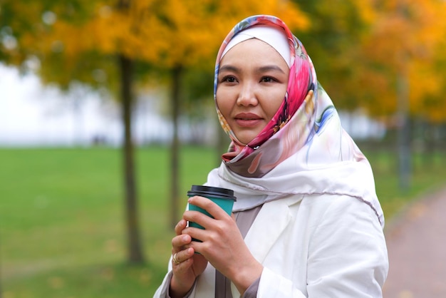 Retrato de hermosa mujer joven musulmana bastante asiática, niña islámica lleva en su cabeza pañuelo hijab y sosteniendo una taza de té o café en la mano, sonriendo. Ropa árabe musulmana tradicional.