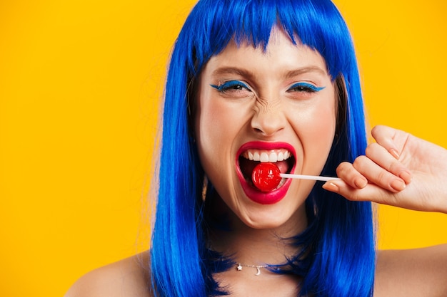 Retrato de hermosa mujer joven con peluca azul comiendo piruletas y mirando aislado sobre pared amarilla