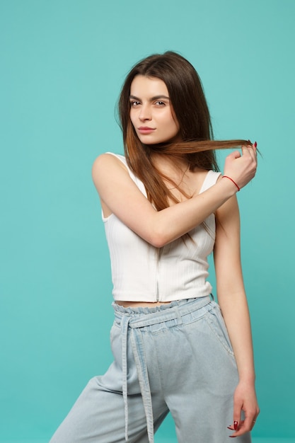 Retrato de hermosa mujer joven en ropa casual ligera mirando a cámara, sosteniendo el cabello aislado sobre fondo azul turquesa en estudio. Personas sinceras emociones, concepto de estilo de vida. Simulacros de espacio de copia.