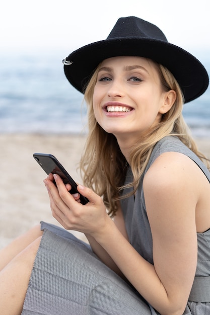 Retrato de hermosa mujer joven rubia sosteniendo un teléfono móvil y mirando a la cámara con una sonrisa perfecta con un sombrero en la playa