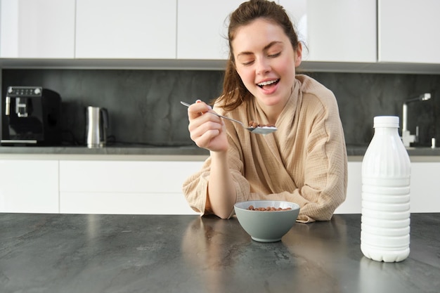 Retrato de una hermosa mujer joven y saludable en bata de baño desayuna en la cocina tiene cereales