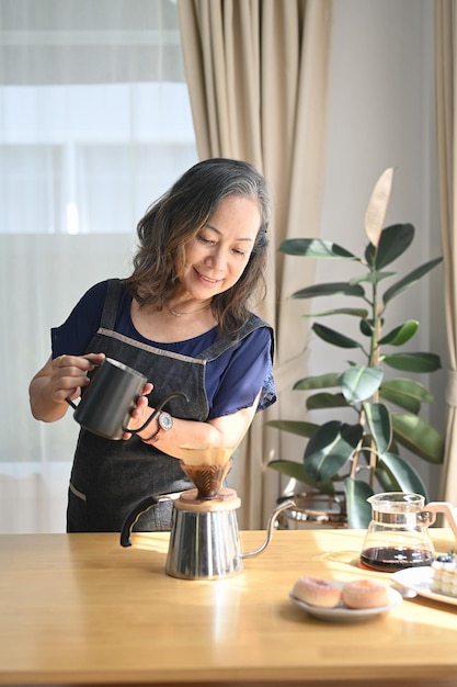 Retrato de una hermosa mujer jubilada que prepara café por goteo y disfruta de su mañana en la mesa de la cocina