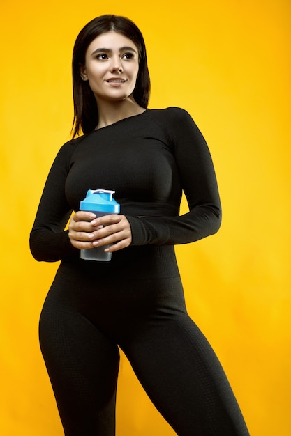 Retrato de una hermosa mujer latina positiva del cuerpo en un traje deportivo negro con una botella de agua posando en amarillo