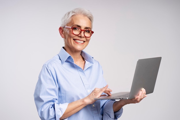 Retrato de una hermosa mujer madura sonriente con elegantes gafas rojas usando una computadora portátil mirando a la cámara aislada en un fondo gris Mujer de negocios de cabello gris concepto de negocio exitoso