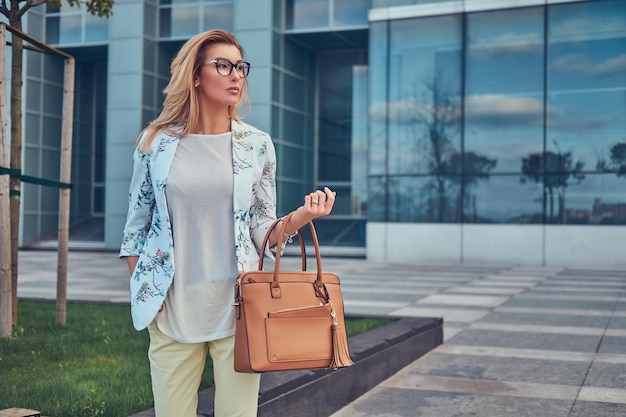 Retrato de una hermosa mujer de moda con ropa elegante y gafas con un bolso, parada contra un rascacielos.