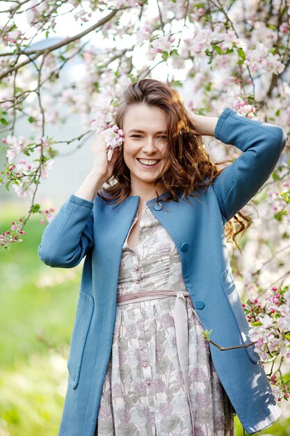 Retrato de una hermosa mujer morena en chaqueta azul en jardín de manzano en flor en primavera. Disfruta de la naturaleza. Niña sonriente sana al aire libre. Concepto de primavera. Hermosa chica en huerto