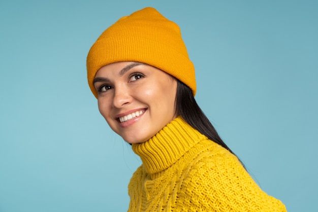 Retrato de hermosa mujer morena joven con ropa de invierno abrigada mirando con alegría a la cámara. Chica guapa con gorro de punto y chaqueta aislado sobre fondo azul.