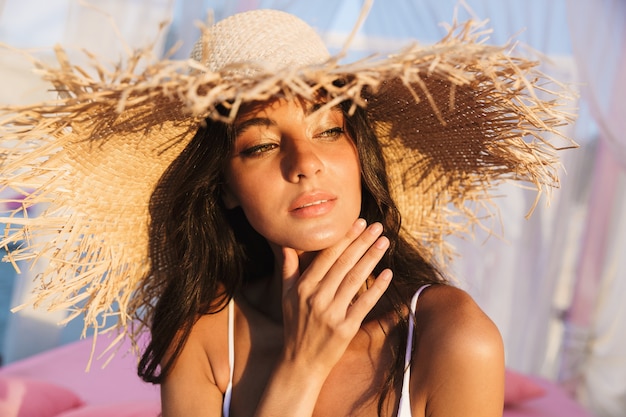 Retrato de hermosa mujer morena en traje de baño y sombrero de paja sentada en la cama de la playa lounge en la mañana en el resort de verano