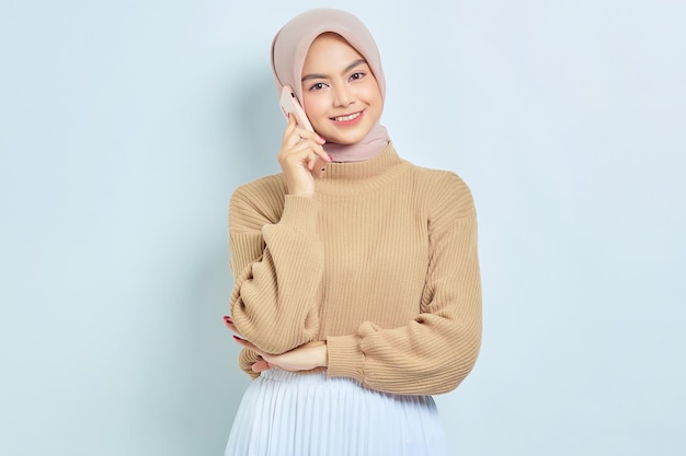 Retrato de una hermosa mujer musulmana asiática sonriente con suéter marrón e hiyab hablando por teléfono móvil aislado sobre fondo blanco Concepto de estilo de vida religioso de la gente