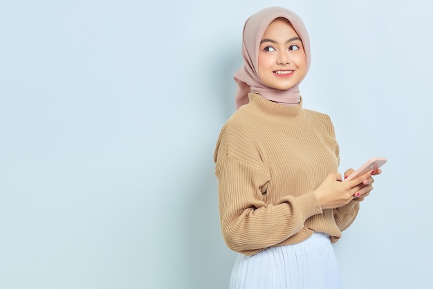 Retrato de una hermosa mujer musulmana asiática sonriente con suéter marrón e hiyab usando un teléfono móvil y mirando a un lado aislado sobre fondo blanco Concepto de estilo de vida religioso de la gente