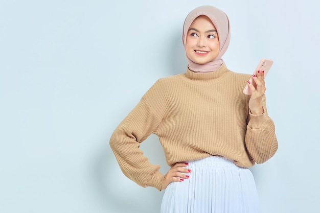 Retrato de una hermosa mujer musulmana asiática sonriente con suéter marrón y hiyab usando un teléfono móvil mirando hacia el espacio en blanco aislado sobre fondo blanco Concepto de estilo de vida religioso de la gente