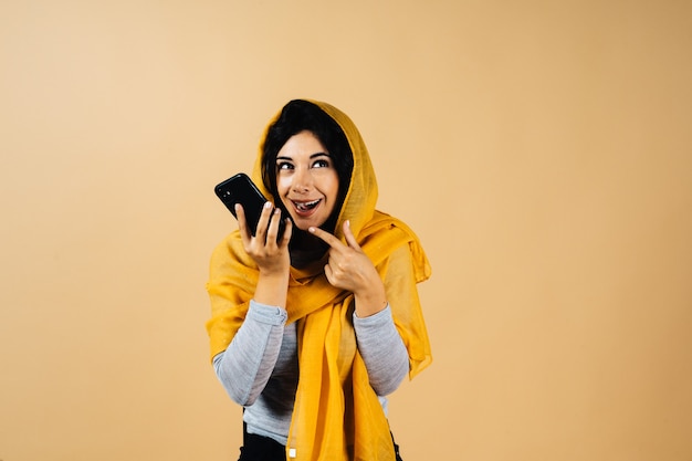 Retrato de hermosa mujer musulmana contra el fondo naranja usando su celular para finalizar un mensaje de audio