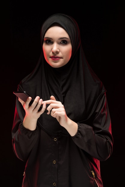 Retrato de hermosa mujer musulmana joven amigable positiva vistiendo hijab negro con teléfono móvil sobre fondo negro