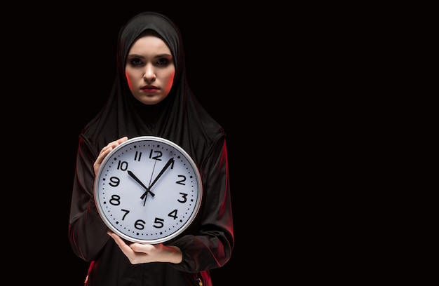 Retrato de hermosa mujer musulmana joven asustada seria asustada vistiendo hijab negro con reloj en sus manos como el tiempo se acaba el concepto sobre fondo negro