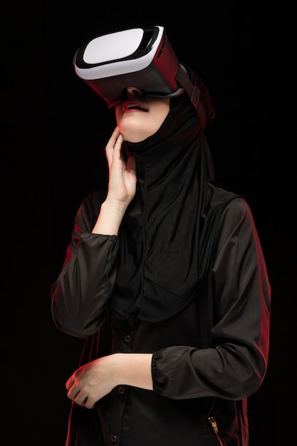 Retrato de hermosa mujer musulmana joven inteligente vistiendo hijab negro con casco de realidad virtual sobre fondo negro