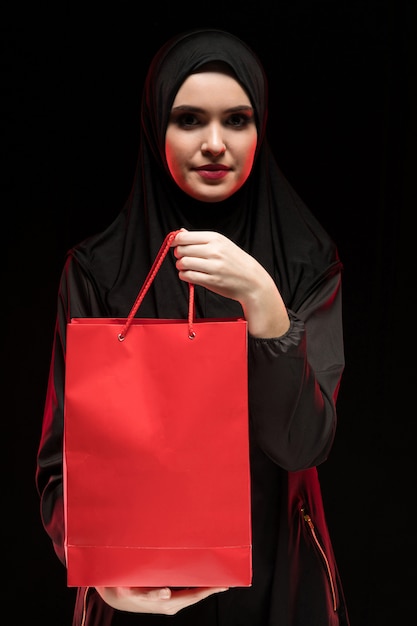 Retrato de hermosa mujer musulmana joven inteligente vistiendo hijab negro ofreciendo bolsa de compras como dependienta