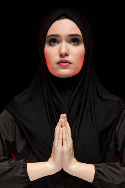 Retrato de hermosa mujer musulmana joven seria con hiyab negro con las manos cerca de su rostro como concepto de oración