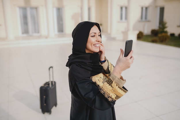 Retrato de hermosa mujer musulmana en ropa tradicional usando un teléfono inteligente para llamar a un taxi mientras está de pie delante de su casa. En el equipaje de fondo.