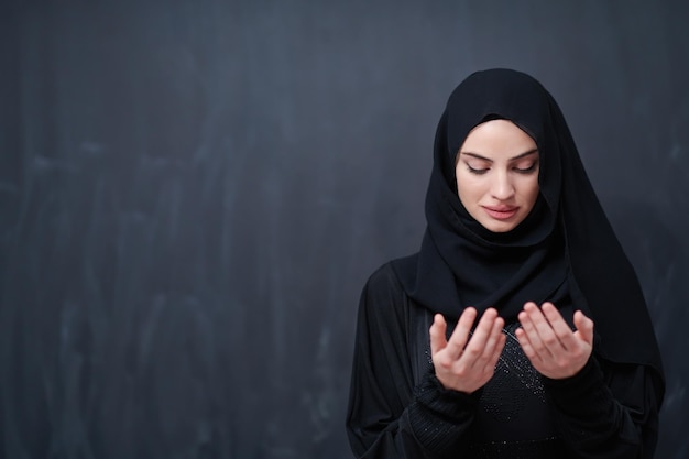 retrato de una hermosa mujer musulmana vestida de moda con hiyab haciendo oración tradicional a Dios mantiene las manos en gesto de oración aislado en un fondo de pizarra negra que representa el islam moderno fas