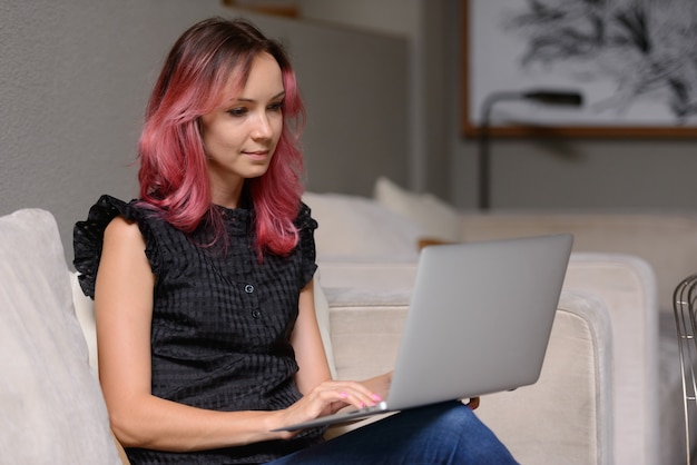 Retrato de hermosa mujer de negocios con cabello rosado usando laptop en la sala de estar como concepto de trabajo desde casa