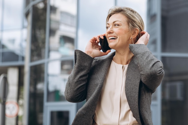 Retrato de una hermosa mujer de negocios madura con traje y chaqueta gris sonriendo y hablando por teléfono en el moderno fondo urbano y de edificios de oficinas
