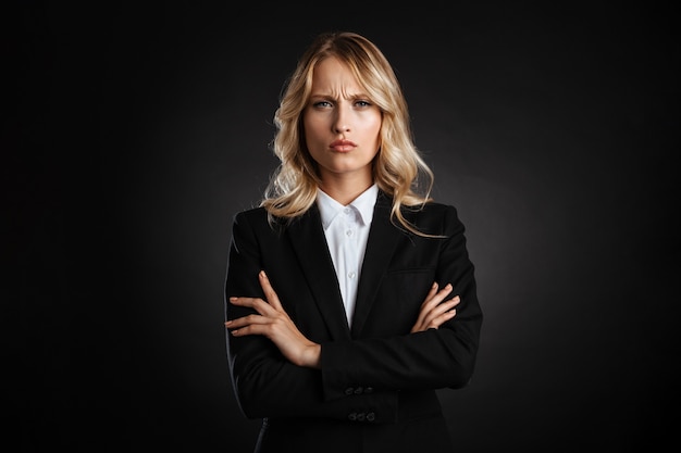 Retrato de una hermosa mujer de negocios de pelo rubio confundido vestida con ropa formal que se encuentran aisladas sobre la pared negra, con los brazos cruzados