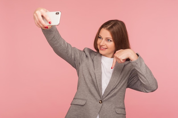 Retrato de una hermosa mujer de negocios positiva con chaqueta de traje haciendo videollamadas y apuntando hacia abajo para suscribirse a una conferencia en línea usando una toma de estudio de comunicación móvil aislada