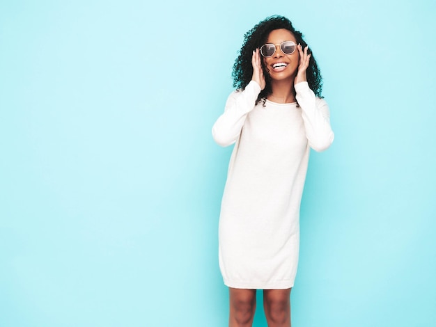 Retrato de hermosa mujer negra con peinado de rizos afro Modelo sonriente vestida con vestido blanco de verano Sexy mujer despreocupada posando junto a la pared azul en el estudio Bronceada y alegre Aislada