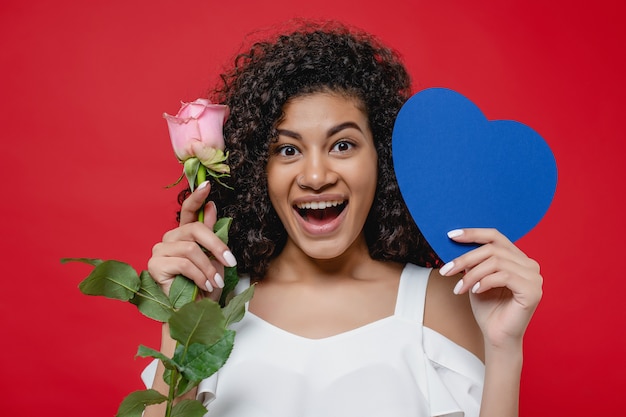 Retrato de hermosa mujer negra sonriente con rosa rosa y tarjeta en forma de corazón azul aislado sobre rojo