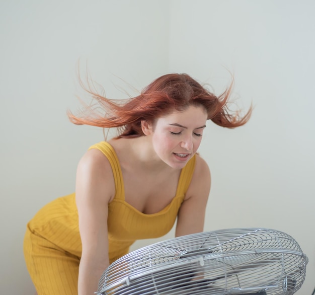Retrato de una hermosa mujer pelirroja con overoles mostaza disfrutando de la brisa refrescante de un gran ventilador eléctrico La niña se refresca en el calor sofocante del verano El cabello se desarrolla a partir del viento