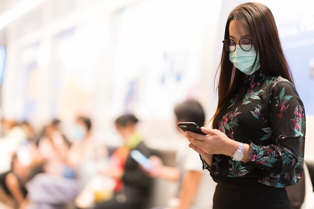 Retrato Una hermosa mujer de pie con un teléfono móvil para consultar el correo electrónico y usar una mascarilla con fondo borroso Bokeh